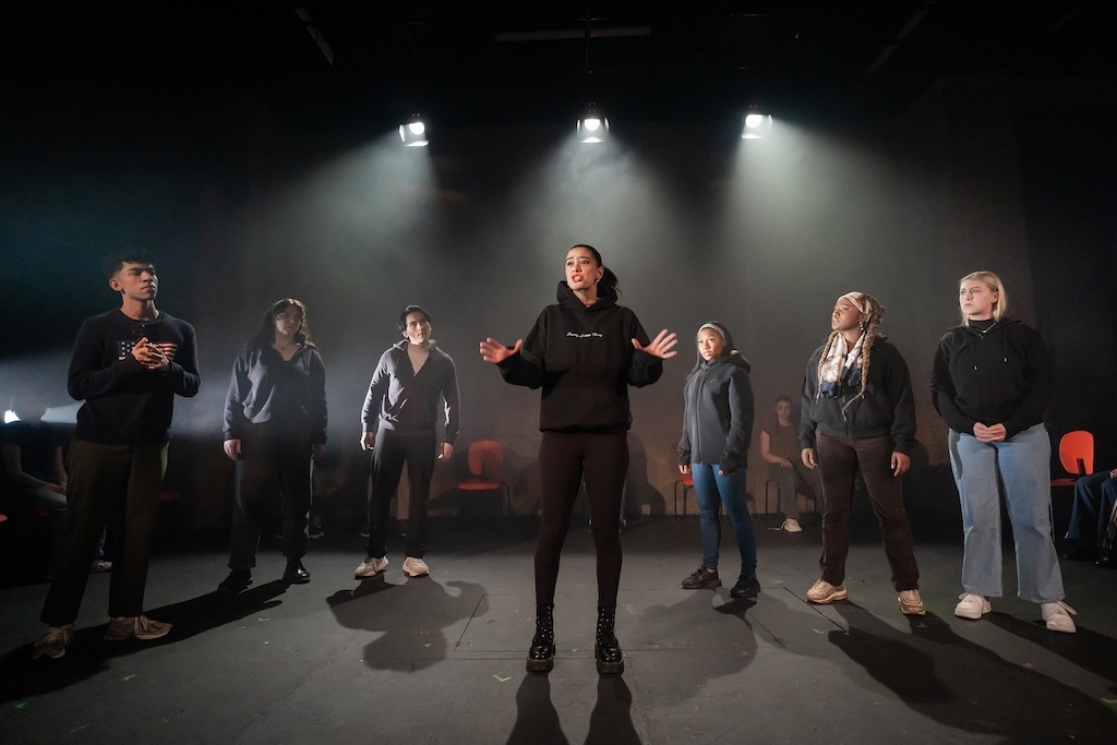 Action shot of the spring 2024 Shakespeare in Performance at RADA final presentation of Julius Caesar. An actor stands in the middle of the stage, in mid-performance, delivering a monologue. Six additional actors stand in a semicircle around her.